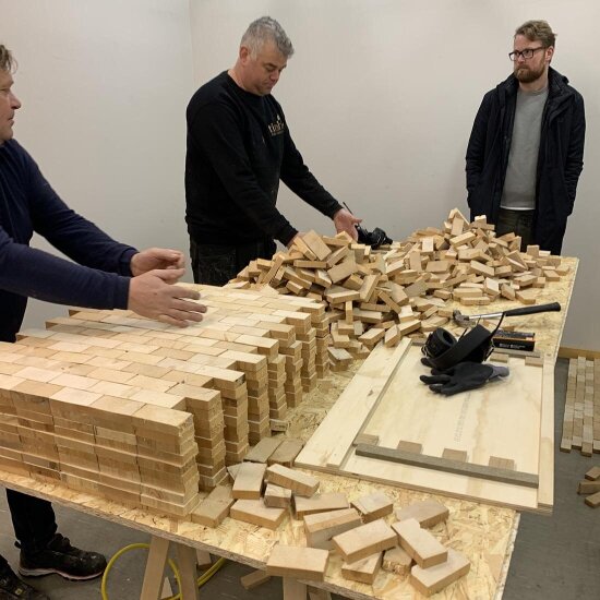 Tre menn rundt et bord fullt av trematerialer fra avkapp.